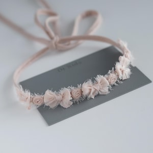 May include: A pink fabric headband with a row of small fabric flowers and bows. The headband is tied with a long ribbon.