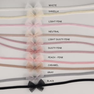 Può includere: Una selezione di fasce per neonati in vari colori, tra cui bianco, vaniglia, rosa chiaro, neutro, rosa chiaro polveroso, rosa polveroso, pesca-rosa, caramello, grigio e nero. Ogni fascia presenta un delicato fiocco.