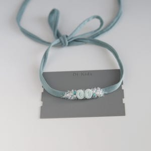 Op de afbeelding: Een lichtblauw hoofdband met een witte en lichtblauwe bloemenversiering. Het hoofdband is vastgebonden met een lichtblauw lint. De tekst "Di Kids" is gedrukt op een grijze kaart achter het hoofdband.