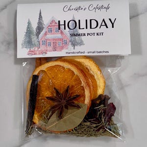 May include: A clear plastic bag containing a holiday simmer pot kit. The kit includes dried orange slices, a star anise, a cinnamon stick, rosemary, and other spices. The bag is topped with a label that reads "Christie's Celestials HOLIDAY Simmer Pot Kit."