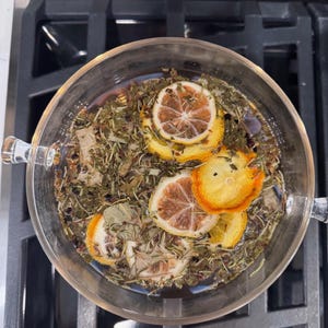 May include: A clear glass bowl filled with herbal tea, featuring dried lemon slices and various herbs. The tea is steeping, with the ingredients visible in the water. The bowl has two handles and sits on a dark stovetop.