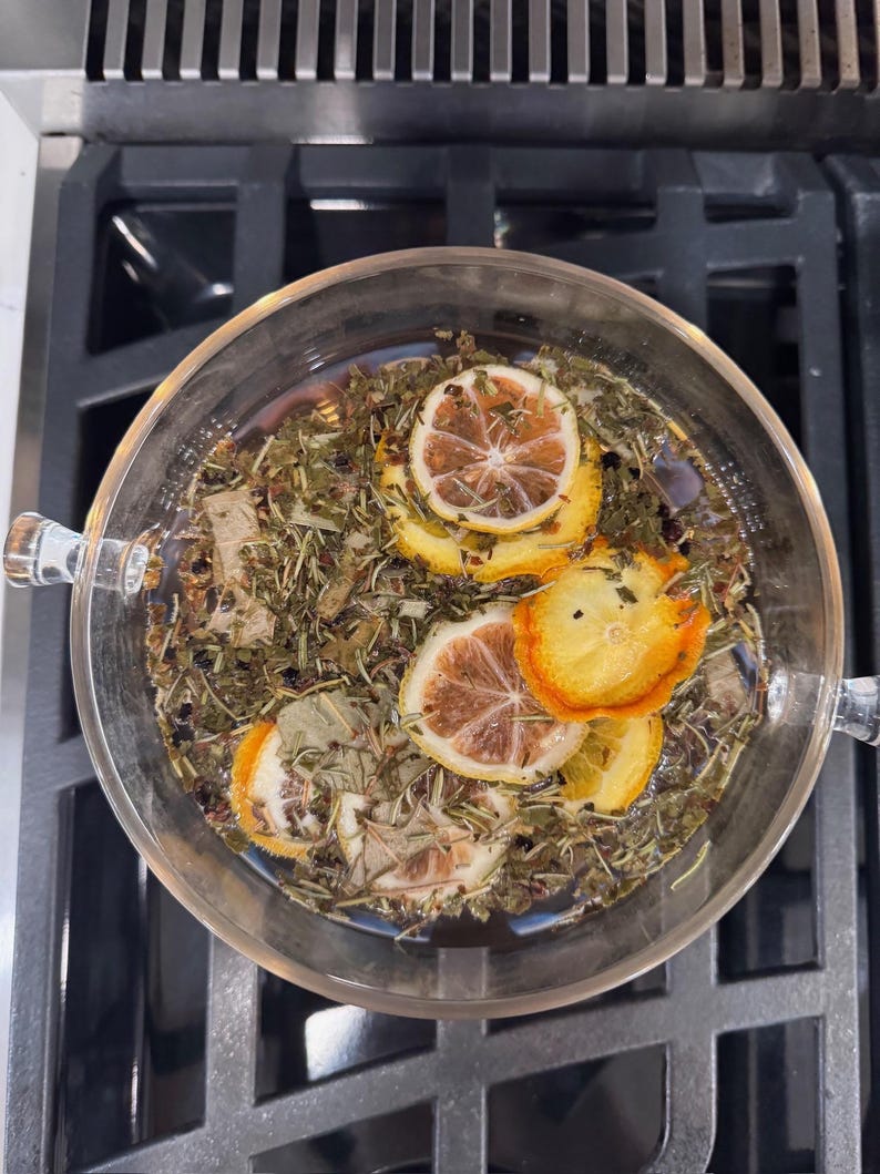 May include: A clear glass bowl filled with herbal tea, featuring dried lemon slices and various herbs. The tea is steeping, with the ingredients visible in the water. The bowl has two handles and sits on a dark stovetop.