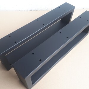 Könnte beinhalten: Zwei schwarze Metall-Tischbeine mit rechteckigem Design. Jedes Bein hat eine hohle rechteckige Form mit vorgebohrten Löchern zur Montage. Die Beine sind aus mattschwarzem Metall gefertigt und für den Möbelbau konzipiert.