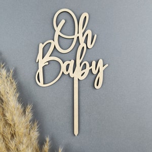 May include: Wooden cake topper with the words "Oh Baby" in a cursive font. The topper is on a gray background.