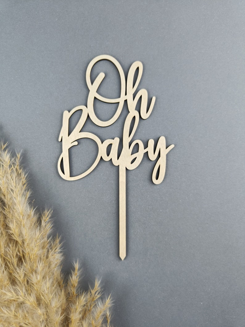 May include: Wooden cake topper with the words "Oh Baby" in a cursive font. The topper is on a gray background.