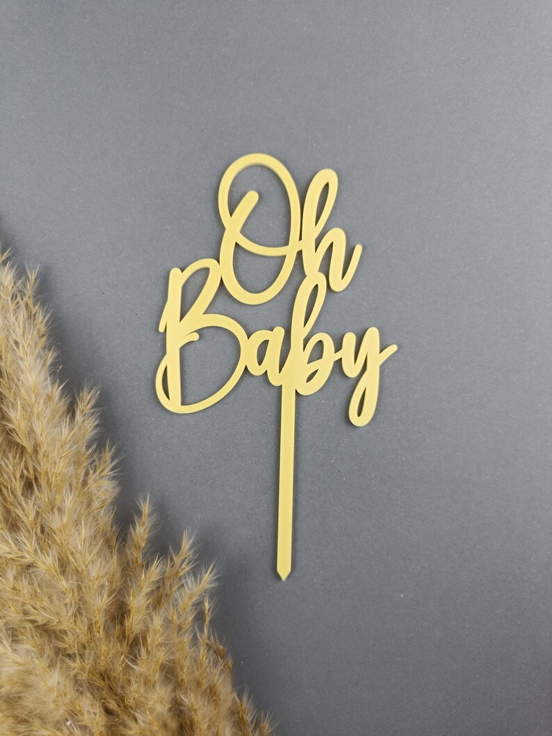 May include: A wooden cake topper with the words "Oh Baby" in a cursive font. The topper is a light brown color and is perfect for a baby shower or gender reveal party.