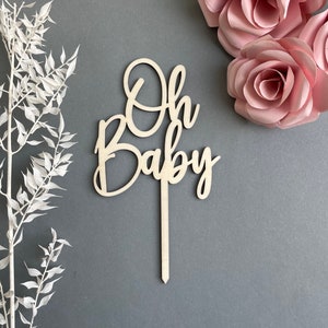 May include: Wooden cake topper with the words "Oh Baby" in a cursive font. The topper is on a gray background with pink paper flowers.