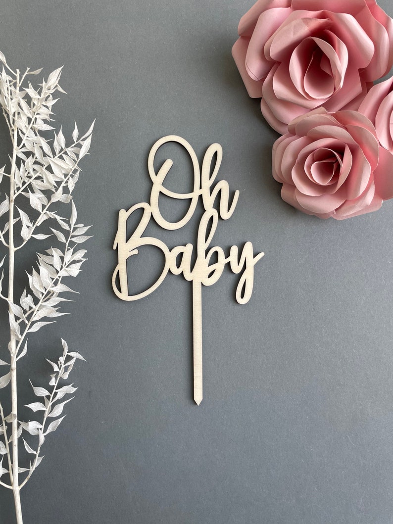 May include: Wooden cake topper with the words "Oh Baby" in a cursive font. The topper is on a gray background with pink paper flowers.