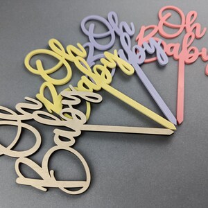 May include: Four wooden cake toppers with the words "Oh Baby" in different colors. The toppers are in shades of pink, purple, yellow, and natural wood.