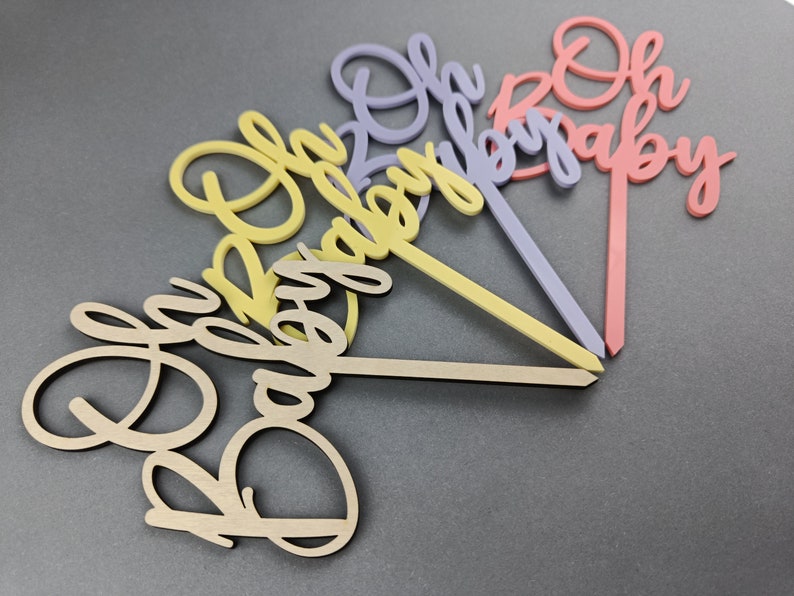 May include: Four wooden cake toppers with the words "Oh Baby" in different colors. The toppers are in shades of pink, purple, yellow, and natural wood.