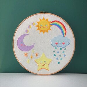 Può includere: Un cerchio da ricamo bianco con un disegno a punto croce colorato che presenta un sole sorridente, un arcobaleno, una luna crescente, una stella e una nuvola con gocce di pioggia.