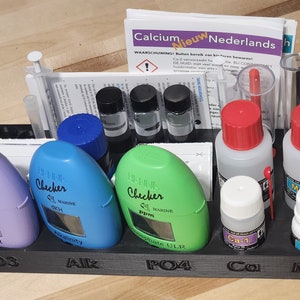 May include: A black plastic test kit organizer with six compartments. The compartments are labeled NO3, Alk, PO4, Ca, and Mg. The organizer contains various test kits, including a purple test kit labeled "Checker Marine OKH Salinity", a blue test kit labeled "Checker Marine dKH", a green test kit labeled "Checker Marine Ppm Phosphate ULTR", a clear bottle labeled "Ca-1", and a clear bottle labeled "Mg-1".