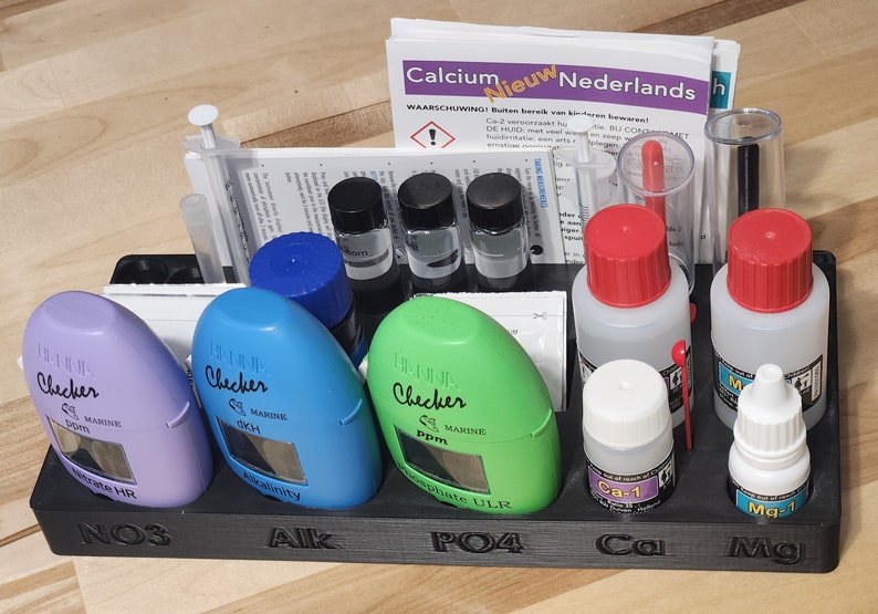 May include: A black plastic test kit organizer with six compartments. The compartments are labeled NO3, Alk, PO4, Ca, and Mg. The organizer contains various test kits, including a purple test kit labeled "Checker Marine OKH Salinity", a blue test kit labeled "Checker Marine dKH", a green test kit labeled "Checker Marine Ppm Phosphate ULTR", a clear bottle labeled "Ca-1", and a clear bottle labeled "Mg-1".
