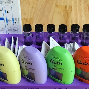 May include: A purple plastic test kit tray with eight Hanna Checker colorimeters for measuring water quality. The colorimeters are labeled with the parameters they measure, including pH, ORP, Salinity, and Magnesium. The tray also includes eight small bottles of reagent solution.