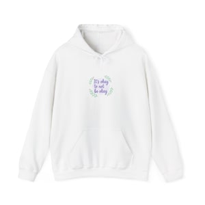Pode incluir: Moletom branco com bolso frontal e capuz com cordão. O moletom apresenta o texto bordado "It's okay to not be okay" em um círculo de folhas verdes e texto roxo.