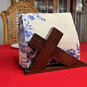 May include: A wooden napkin holder with a dark brown finish holds a stack of white napkins. The napkins are decorated with a blue floral pattern. The holder sits on a red tablecloth.