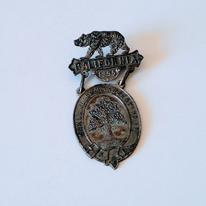May include: A silver-toned metal badge featuring a bear walking on a banner inscribed with "CALIFORNIA 1895". The banner is connected to a larger oval-shaped badge with the text "OAKLAND COMMANDERY NO. 11" and a tree in the centre.