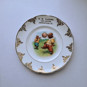 May include: White porcelain plate with gold trim and a scene of two bears playing with a ball. The plate has the text "O. H. Danner, The Leader, Lincolnville, Pa." printed on it.
