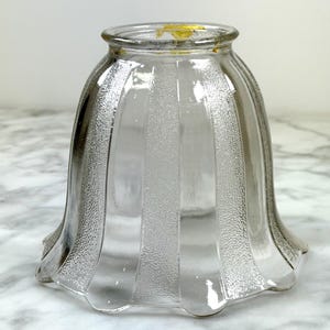 May include: Clear glass lampshade with vertical textured stripes. The lampshade has a flared, scalloped bottom and a rounded top. The glass has a slight tint of grey and yellow.