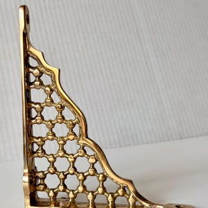 Op de afbeelding: Een goudkleurige hoekbeugel met een sierlijk, opengewerkt ontwerp. De beugel heeft een decoratieve rand en een reeks met elkaar verbonden cirkels die een rasterpatroon vormen. Het heeft een montagegat voor een veilige installatie. De algehele esthetiek is vintage of antiek.