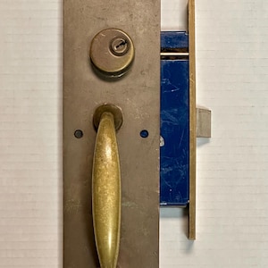 Może przedstawiać: Zestaw zabytkowych klamek do drzwi z prostokątną, zmatowioną metalową płytą. Posiada mosiężną klamkę, otwór na klucz i niebieski mechanizm zatrzaskowy. Okucia są przeznaczone do drzwi.