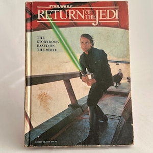 May include: A vintage copy of "Star Wars: Return of the Jedi". The book cover displays Luke Skywalker wielding a green lightsaber. The title is in red and white, with the text "The Storybook Based on the Movie". Published by Weekly Reader Books.