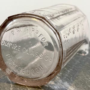 Puede incluir: Recipiente de medición de vidrio transparente con base octogonal. El texto "PAT APPD FOR" y "UMPIRE GLASS CO PITTSBURGH" son visibles en la base. El recipiente tiene marcas de medición en el lateral.