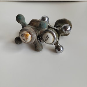May include: Two vintage faucet handles. One handle has a white face with a smiley face, the other has a white face with the word "HOT". The handles are made of metal with a tarnished finish and green oxidation.
