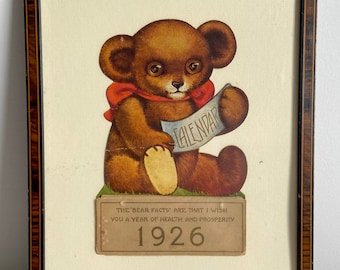 May include: A framed vintage calendar featuring a brown teddy bear holding a scroll with the word "CALENDAR" printed on it. The calendar is from 1926 and includes the text "The 'bear facts' are that I wish you a year of health and prosperity."