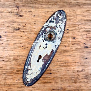 May include: An oval metal door plate with a distressed white painted finish over a dark bronze base. The plate features an embossed decorative border, a central circular opening for a knob, and a lower keyhole opening with two screw holes.