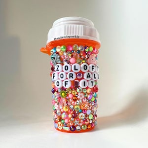 May include: An orange pill bottle decorated with colorful beads, gems, and charms. The bottle has the text "ZOLOFT FOR ALL OF IT" spelled out in white letter beads. The top of the bottle is white.