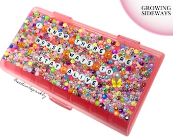 Puede incluir: Un estuche rectangular de plástico rosa decorado con una densa disposición de cuentas coloridas, pedrería y un dije de mariposa amarilla. Cuentas blancas con letras forman la frase I KNOW THERE ARE WORSE WAYS TO STAY ALIVE en la superficie.