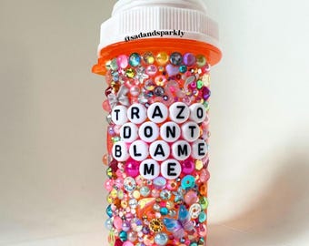Puede incluir: Un frasco de pastillas naranja decorado con una variedad de cuentas de colores, pedrería y detalles de mariposas. La botella presenta el texto "TRAZO DONT BLAME ME" en cuentas de letras blancas. La parte superior de la botella es blanca con el texto "@sadandsparkly".
