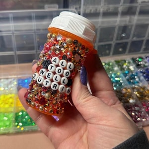 Puede incluir: Un frasco de pastillas naranja adornado con pedrer&iacute;a y cuentas. El frasco tiene las palabras "ALL TOO WELL BUT R IN" escritas con cuentas de letras blancas. El frasco tiene una tapa blanca.