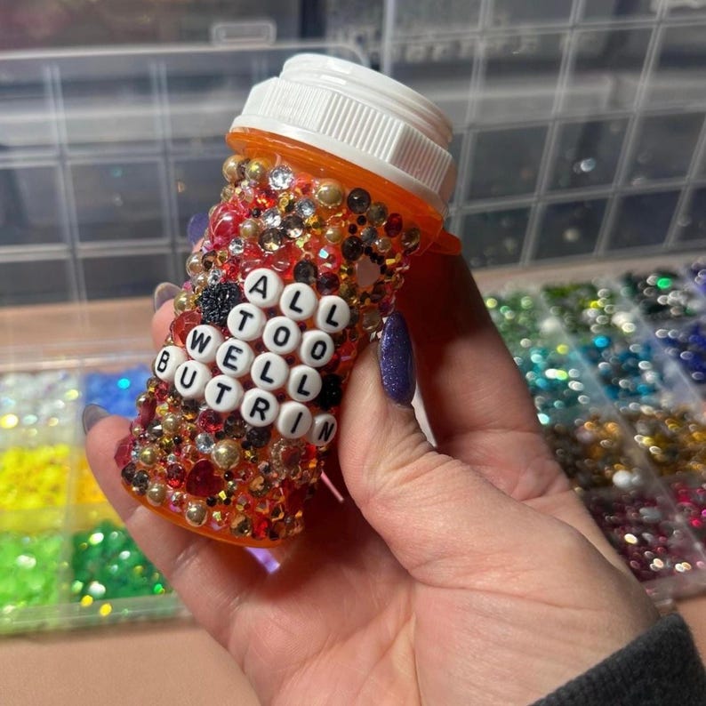 Puede incluir: Un frasco de pastillas naranja adornado con pedrer&iacute;a y cuentas. El frasco tiene las palabras "ALL TOO WELL BUT R IN" escritas con cuentas de letras blancas. El frasco tiene una tapa blanca.