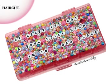 Puede incluir: Un estuche de almacenamiento rectangular de plástico rosa decorado con una densa disposición de cuentas coloridas, pedrería y cuentas de letras blancas que forman la frase: AT LEAST I'VE GOT A SOUL STILL EVEN IF I'M IN A BAD PLACE. El texto @sadandsparkly es visible.