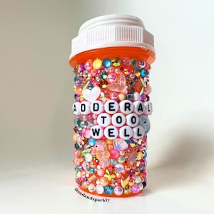 May include: An orange pill bottle decorated with colorful rhinestones, beads, and charms. The bottle has the words "ADDERAL TOO WELL" spelled out in white letter beads. The bottle's base has the text "@sadandsparkly".