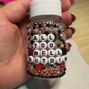 Puede incluir: Una botella de pl&aacute;stico transparente con tapa blanca, adornada con pedrer&iacute;a y cuentas de varios colores. La botella tiene el texto "ALL TOO WELL BUT RIN" escrito con cuentas de letras blancas. Tambi&eacute;n se ven rosas rojas y negras.