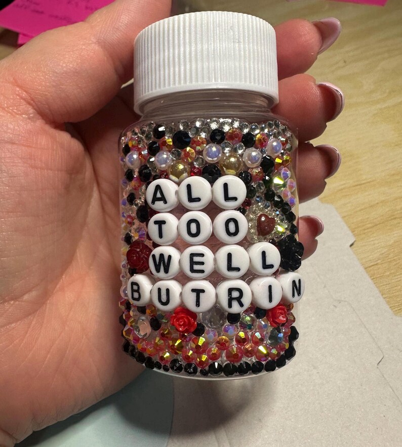 Puede incluir: Una botella de pl&aacute;stico transparente con tapa blanca, adornada con pedrer&iacute;a y cuentas de varios colores. La botella tiene el texto "ALL TOO WELL BUT RIN" escrito con cuentas de letras blancas. Tambi&eacute;n se ven rosas rojas y negras.