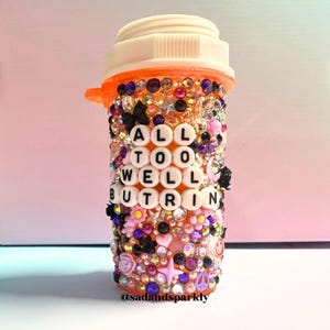 Puede incluir: Un frasco de pastillas naranja, profusamente adornado con brillantes de colores, cuentas y peque&ntilde;os elementos decorativos. El frasco presenta el texto "ALL TOO WELL BUT RIN" en cuentas de letras blancas. El dise&ntilde;o incluye flores, estrellas y un s&iacute;mbolo de la paz.