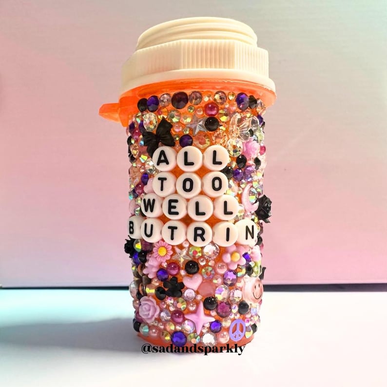 Puede incluir: Un frasco de pastillas naranja, profusamente adornado con brillantes de colores, cuentas y peque&ntilde;os elementos decorativos. El frasco presenta el texto "ALL TOO WELL BUT RIN" en cuentas de letras blancas. El dise&ntilde;o incluye flores, estrellas y un s&iacute;mbolo de la paz.