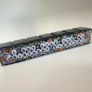 May include: A black, rectangular pill organizer decorated with colorful rhinestones and the phrase "I ACT LIKE IT'S MY BIRTHDAY EVERYDAY" in white letters. The organizer has seven individual compartments with black lids.
