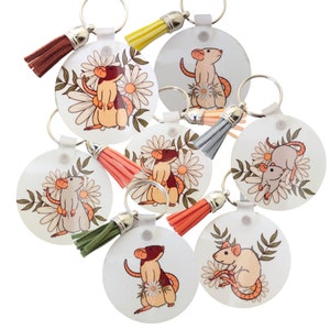 May include: Set of eight keychains featuring a white background with a floral design and a cartoon rat. Each keychain has a different coloured tassel.