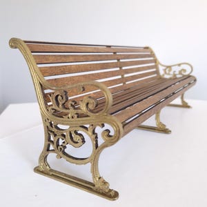 Può includere: Una panchina in legno con una struttura in metallo dorato. La panchina ha un design decorativo sul telaio ed è realizzata con doghe di legno.