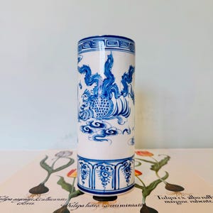 May include: A cylindrical white and blue ceramic vase with a detailed dragon design. The vase features intricate blue patterns and a decorative border. The design includes a dragon, clouds, and floral motifs. The vase is approximately 15 cm tall.