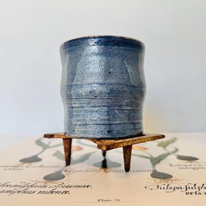 May include: A handmade, blue ceramic cup with a textured surface. The cup sits on a small, square wooden stand with three legs. The cup has a dark blue rim and a lighter blue body.