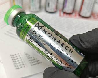 May include: A green glass vial labeled "VITRO SAMPLE" with a silver label that reads "MONARCH" and includes the text "SPECIES: MUTATED ROSE/HUMAN COZILLASAURUS HYBRID", "LOCATION: LAKE ASHI", "NICKNAME: BIOLLANTE", "SAMPLE TYPE: SKIN/CHLOROPHYLL", "LOT: 2009-JUL 011", and "25MG".