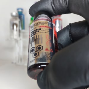 Könnte beinhalten: Eine kleine Glasflasche mit einer roten Flüssigkeit. Die Flasche hat ein Etikett mit der Aufschrift "EXPERIMENTAL TECHNOLOGY PROJECT BLACKLIGHT" und enthält einen Barcode und ein Warnsymbol. Die Flasche wird von einer behandschuhten Hand gehalten.