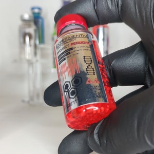 Peut inclure: Une petite bouteille en verre contenant un liquide rouge. La bouteille porte une étiquette argentée avec le texte "EXPERIMENTAL TECHNOLOGY PROJECT REDLIGHT" et d'autres informations. La bouteille est tenue par une main gantée.
