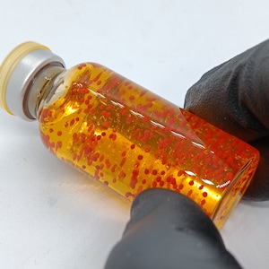 Peut inclure: Une bouteille en verre transparente remplie d'un liquide jaune et de petites perles rouges. La bouteille a un bouchon argenté avec un dessus jaune.
